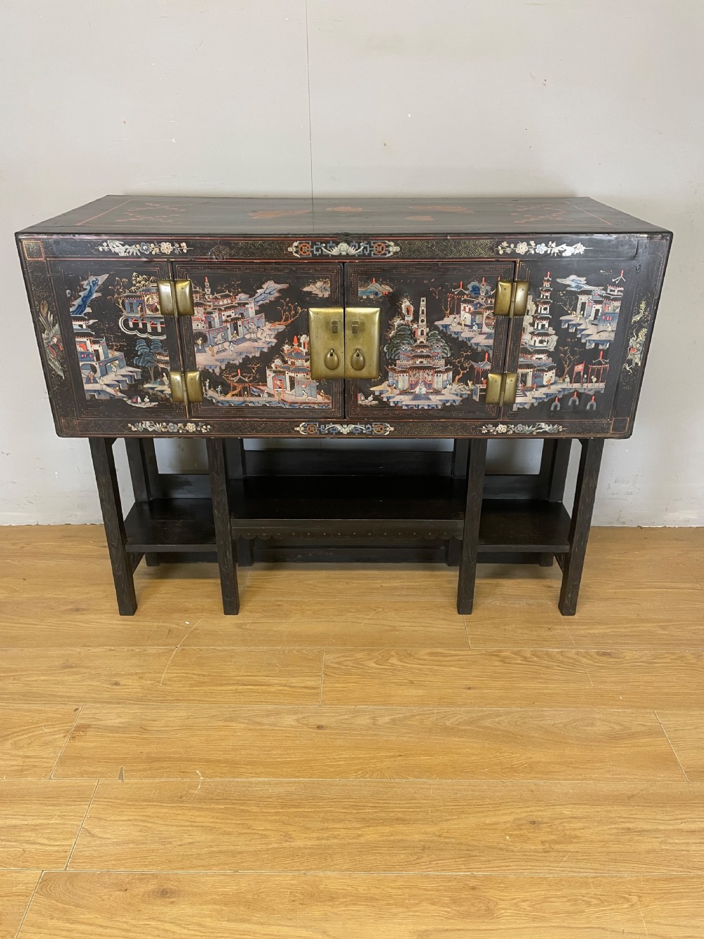 An antique black lacquered cabinet featuring hand-painted landscape scenes and large rectangular brass hinges, elevated on a dark wooden open stand. View II