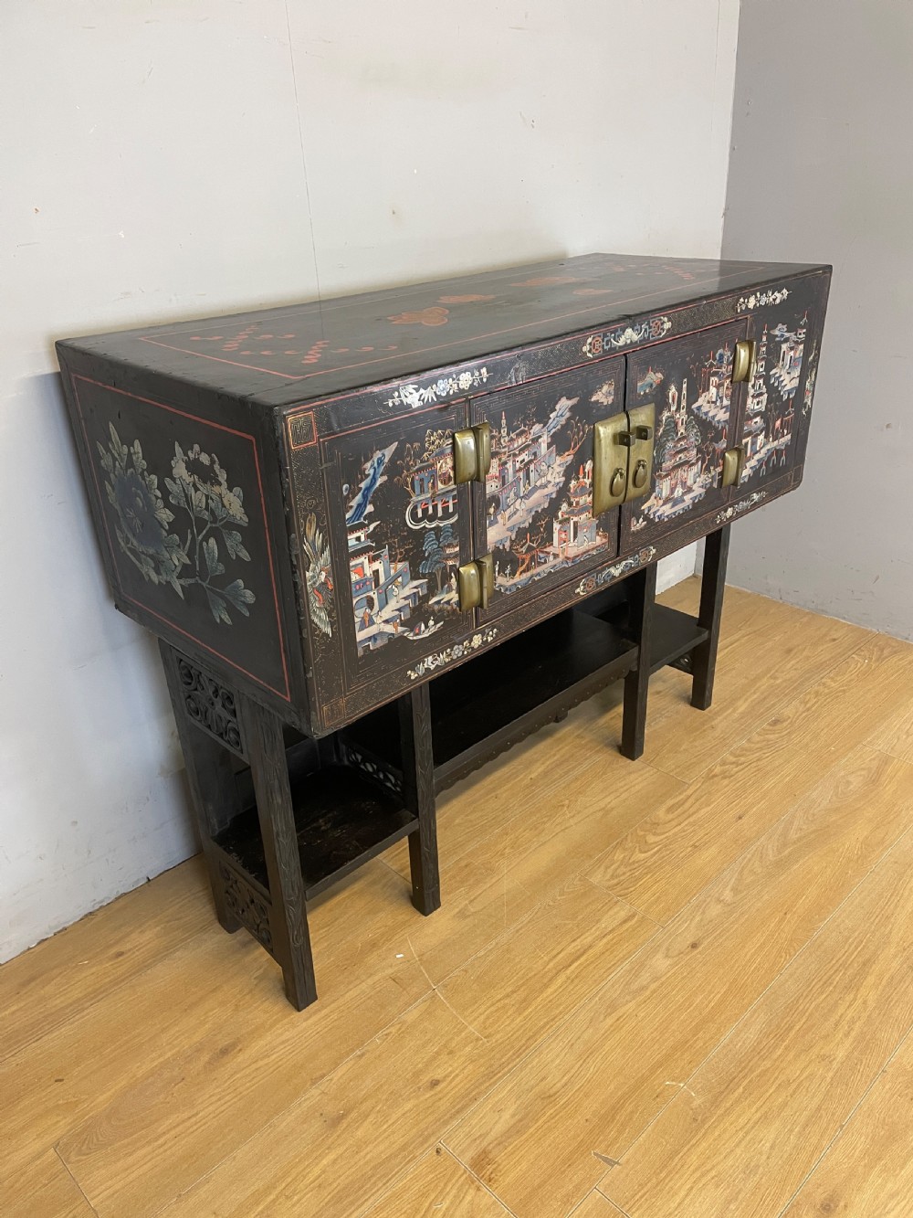 An antique black lacquered cabinet featuring hand-painted landscape scenes and large rectangular brass hinges, elevated on a dark wooden open stand. View III