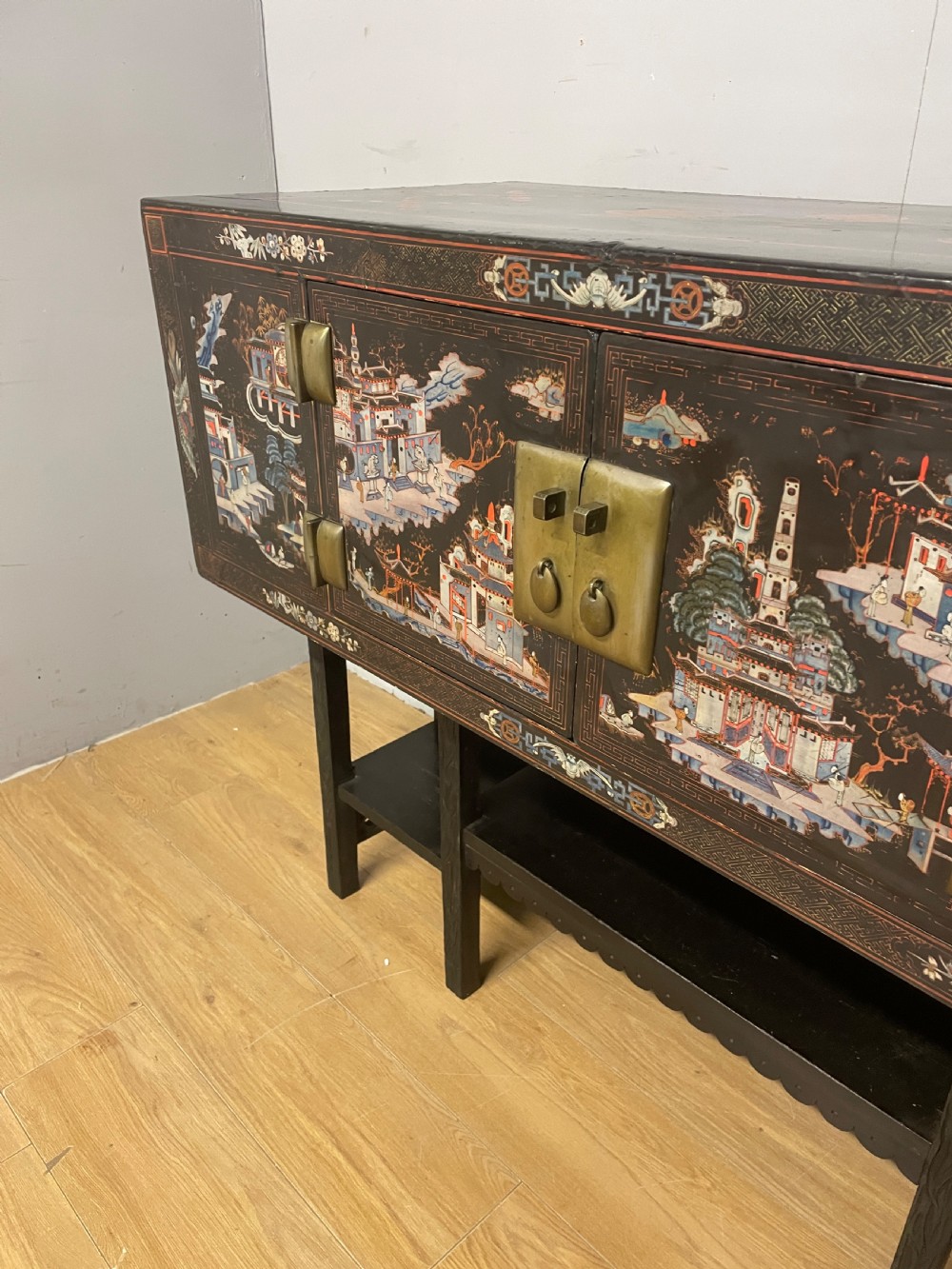 An antique black lacquered cabinet featuring hand-painted landscape scenes and large rectangular brass hinges, elevated on a dark wooden open stand. View VI
