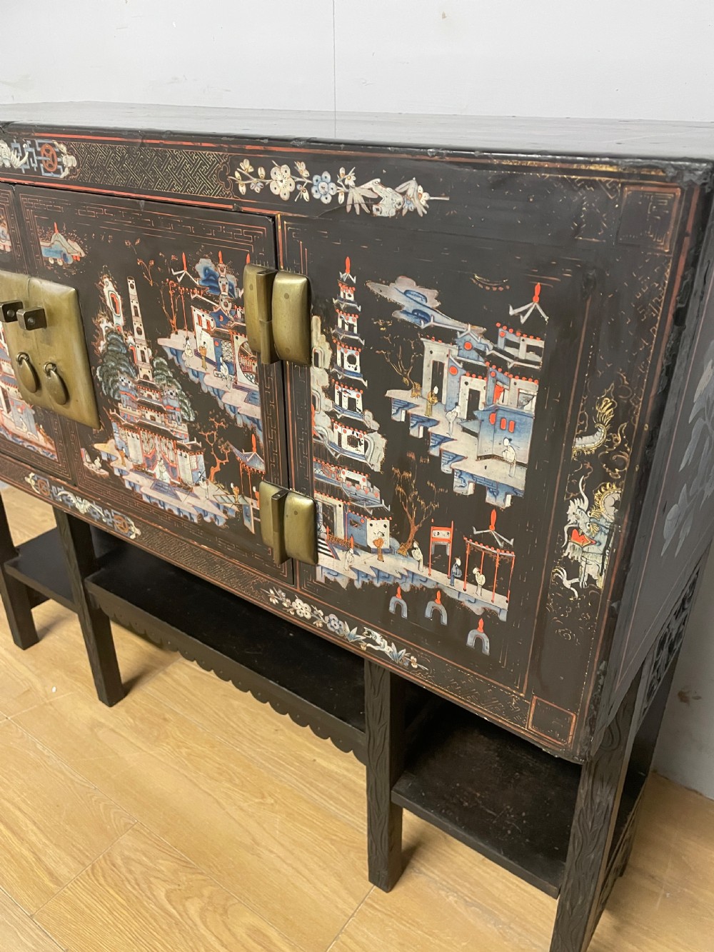 An antique black lacquered cabinet featuring hand-painted landscape scenes and large rectangular brass hinges, elevated on a dark wooden open stand. View VII