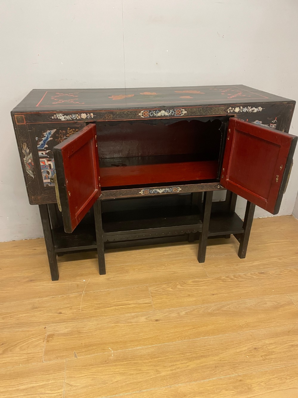 An antique black lacquered cabinet featuring hand-painted landscape scenes and large rectangular brass hinges, elevated on a dark wooden open stand. View X
