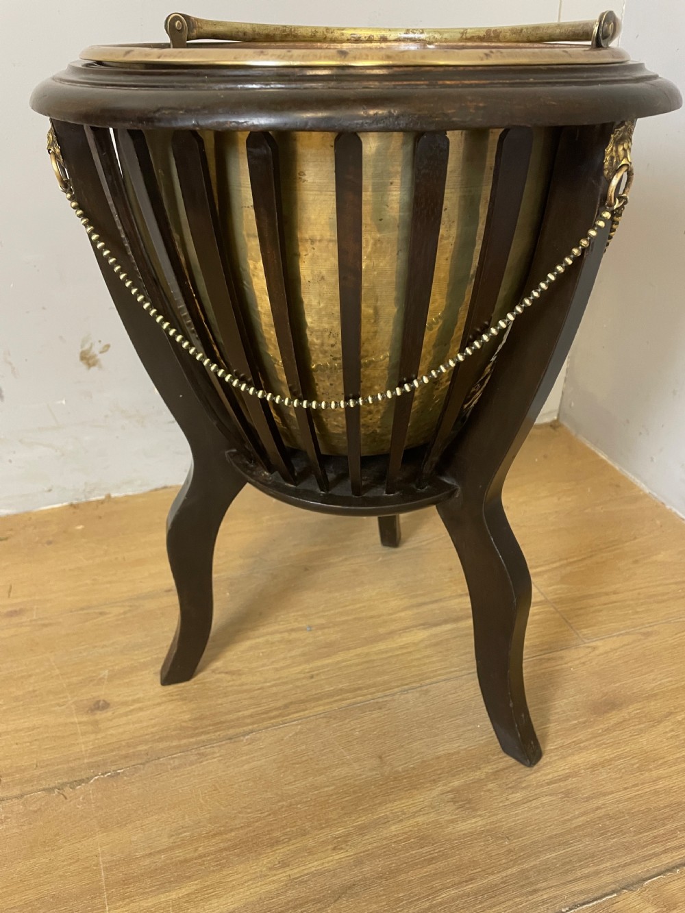 An Edwardian wine cooler featuring a removable brass bucket with a swing handle, supported within a slatted mahogany tripod stand adorned with brass lion mask mounts and decorative chains. View V