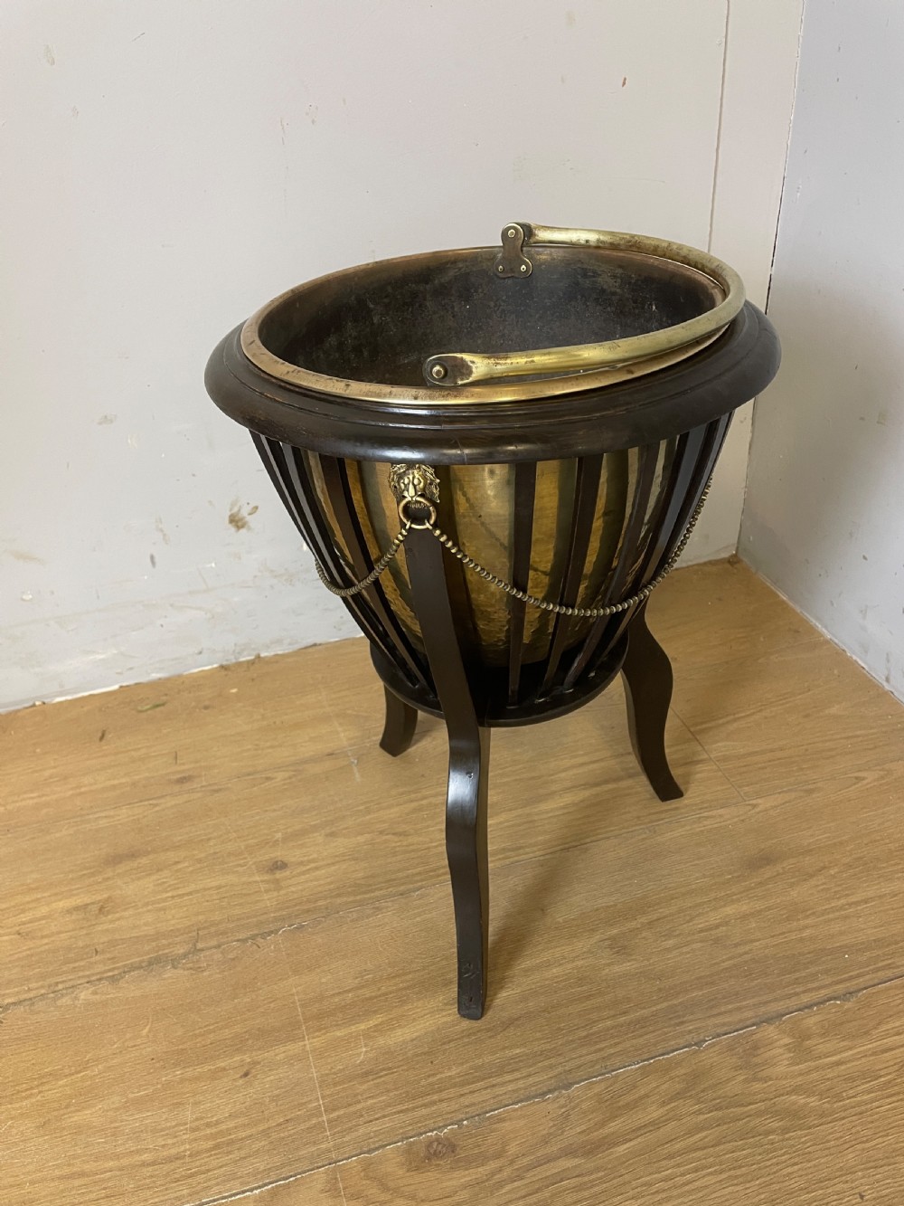 An Edwardian wine cooler featuring a removable brass bucket with a swing handle, supported within a slatted mahogany tripod stand adorned with brass lion mask mounts and decorative chains. View VII
