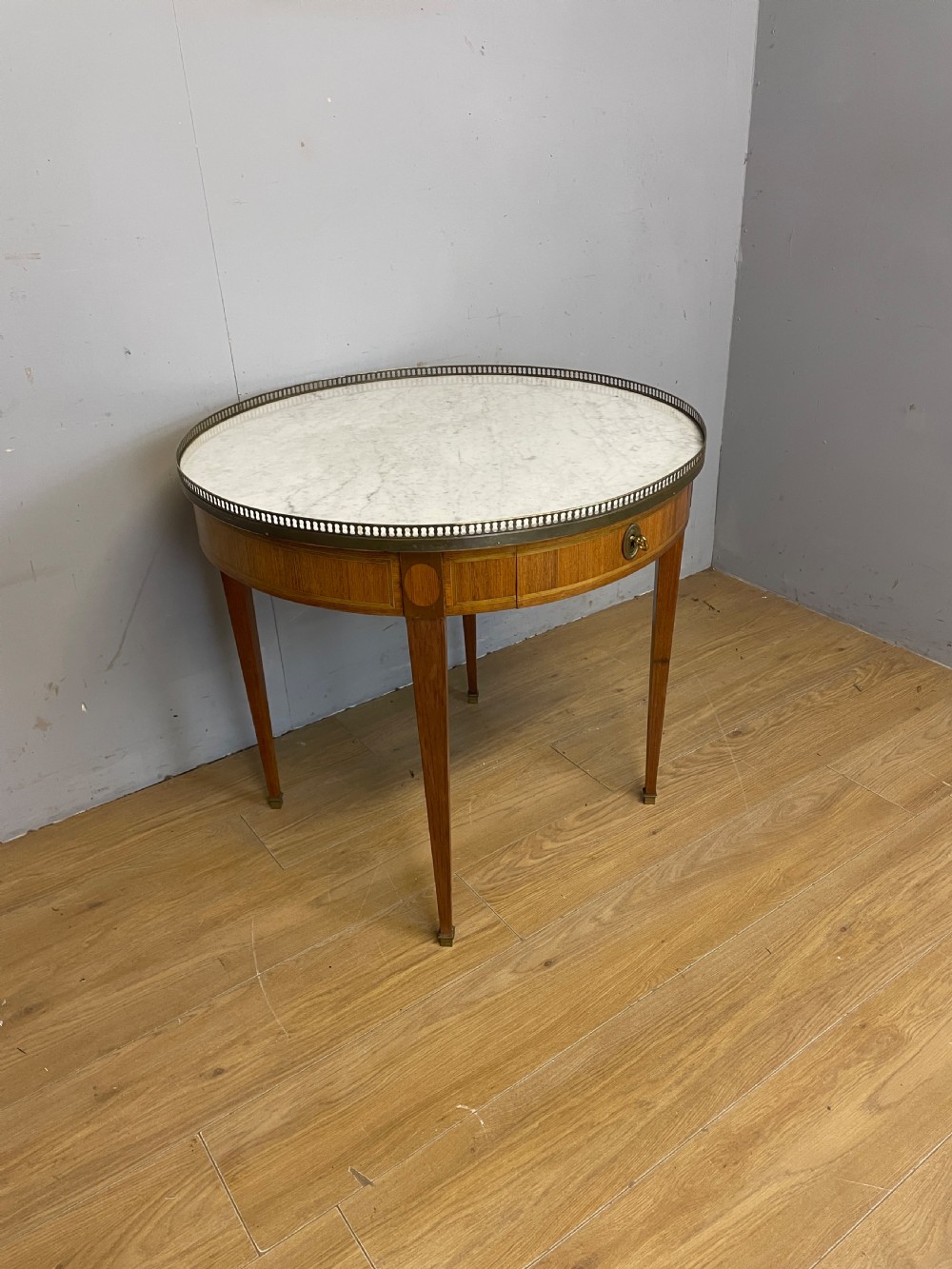 A late 19th-century French circular Bouillotte table featuring a white marble top with a pierced brass gallery, a veneered apron with a small drawer, and four square tapered legs ending in brass sabots.