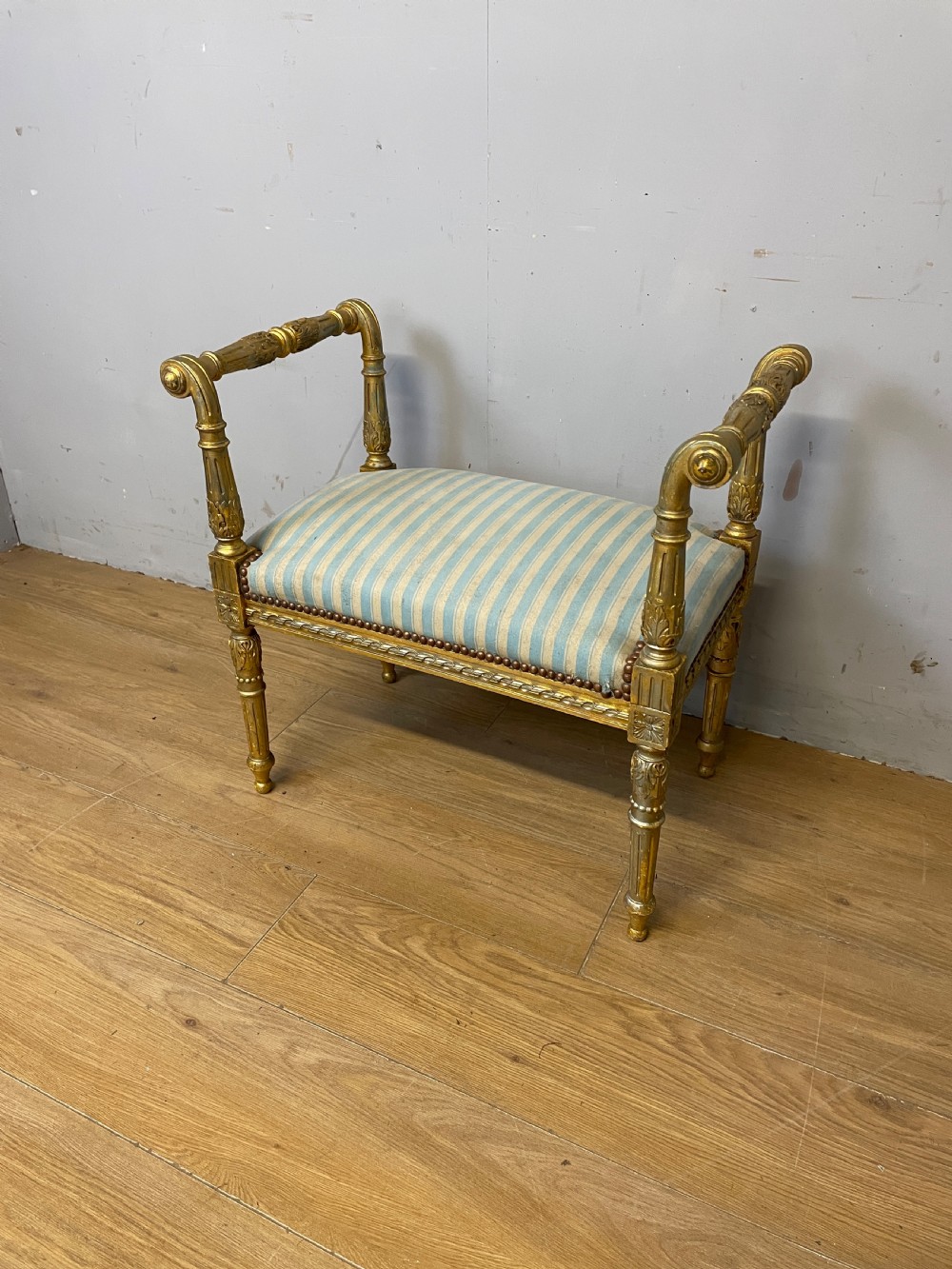A Louis XVI style French giltwood window seat featuring rolled arms, fluted tapering legs, and blue and cream striped upholstery.