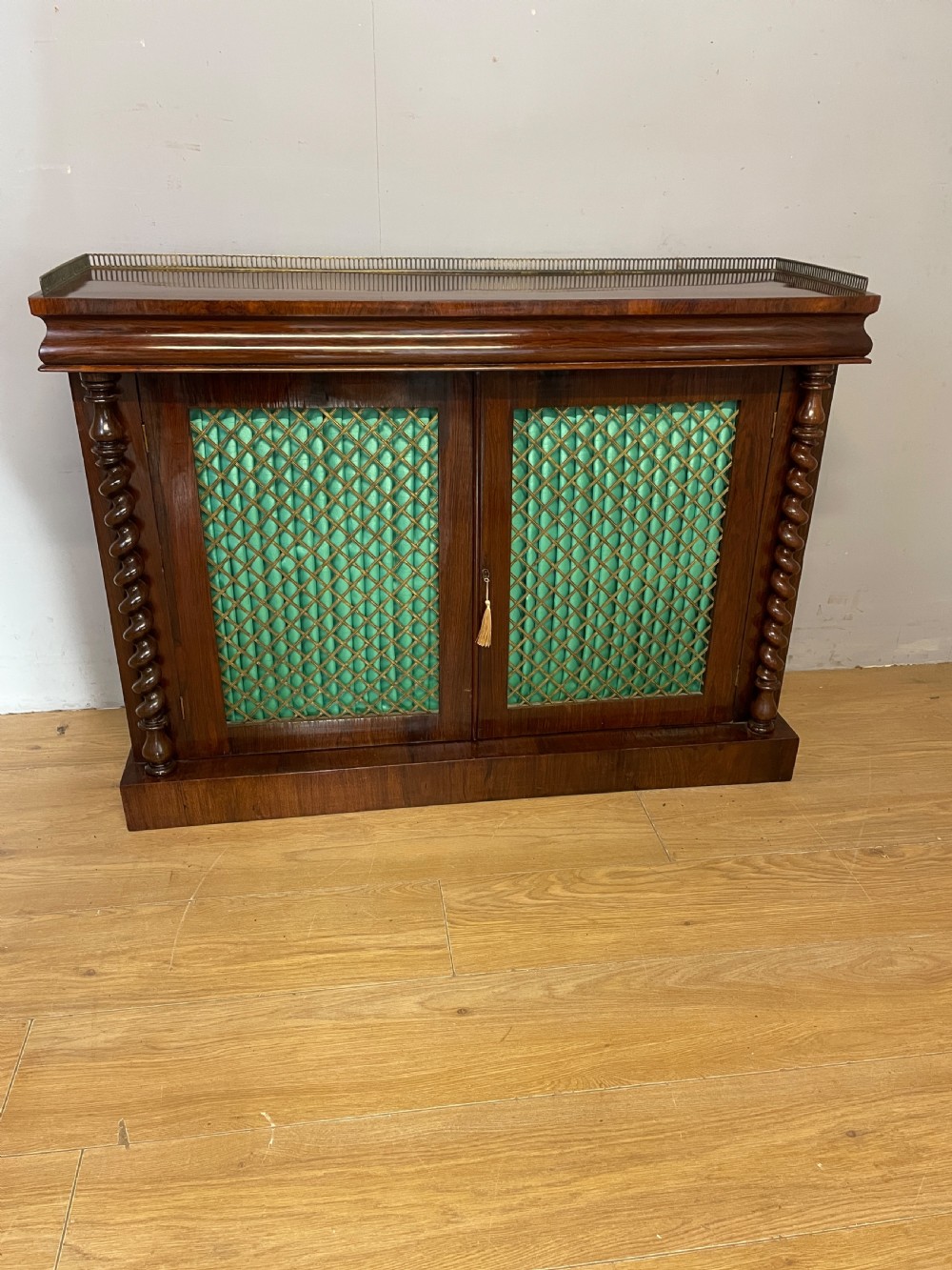 A late Regency rosewood chiffonier featuring barley twist columns, brass wire lattice doors backed with green pleated fabric, and a brass gallery top rail.