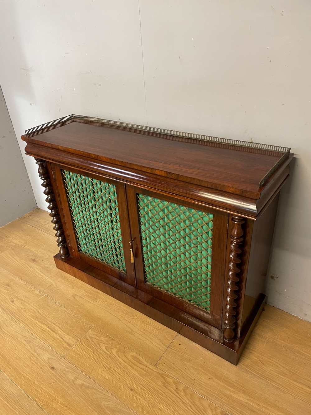A late Regency rosewood chiffonier featuring barley twist columns, brass wire lattice doors backed with green pleated fabric, and a brass gallery top rail. View II