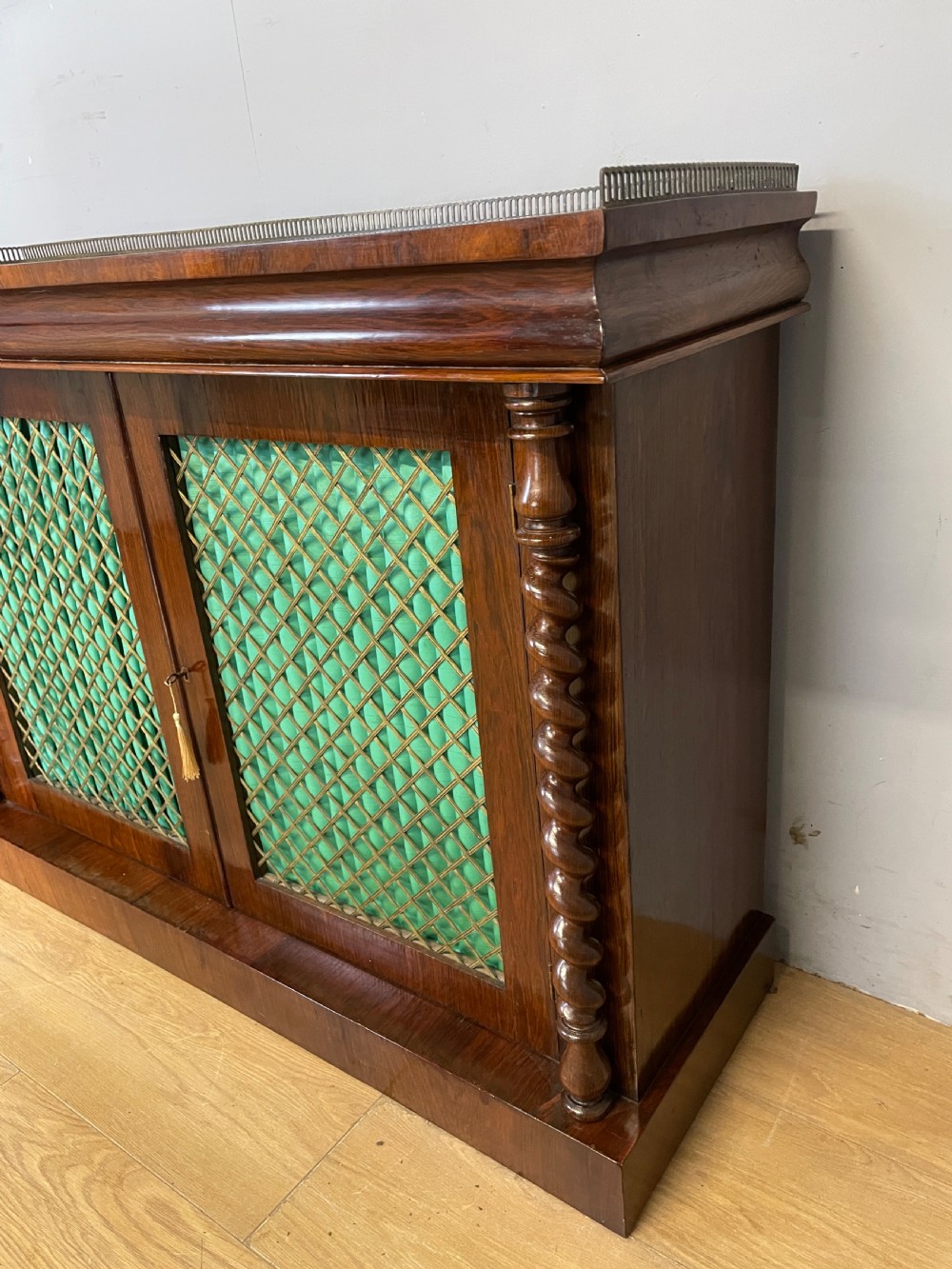 A late Regency rosewood chiffonier featuring barley twist columns, brass wire lattice doors backed with green pleated fabric, and a brass gallery top rail. View VI
