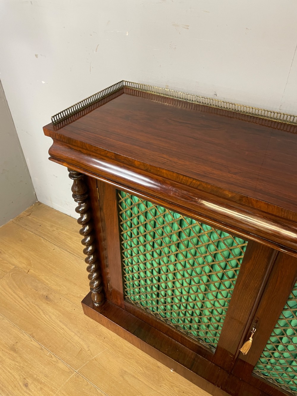 A late Regency rosewood chiffonier featuring barley twist columns, brass wire lattice doors backed with green pleated fabric, and a brass gallery top rail. View X