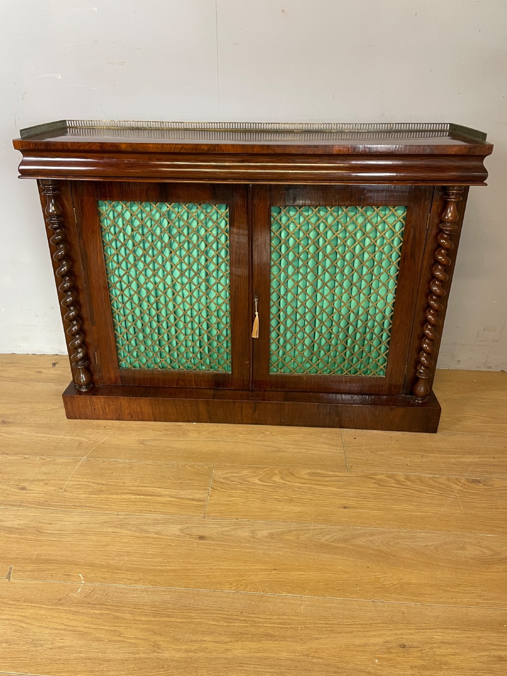 A late Regency rosewood chiffonier featuring barley twist columns, brass wire lattice doors backed with green pleated fabric, and a brass gallery top rail. View XIII