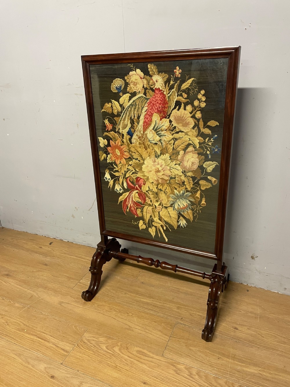 A nineteenth-century mahogany firescreen featuring a central needlepoint panel depicting a floral arrangement and bird, supported by carved splayed legs and a turned stretcher.