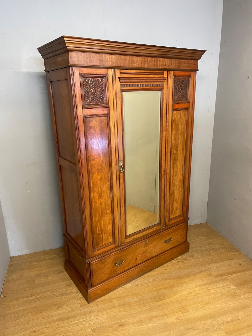 A late 19th-century Arts and Crafts satin walnut wardrobe featuring a central mirrored door, carved foliate panels, and a single base drawer.