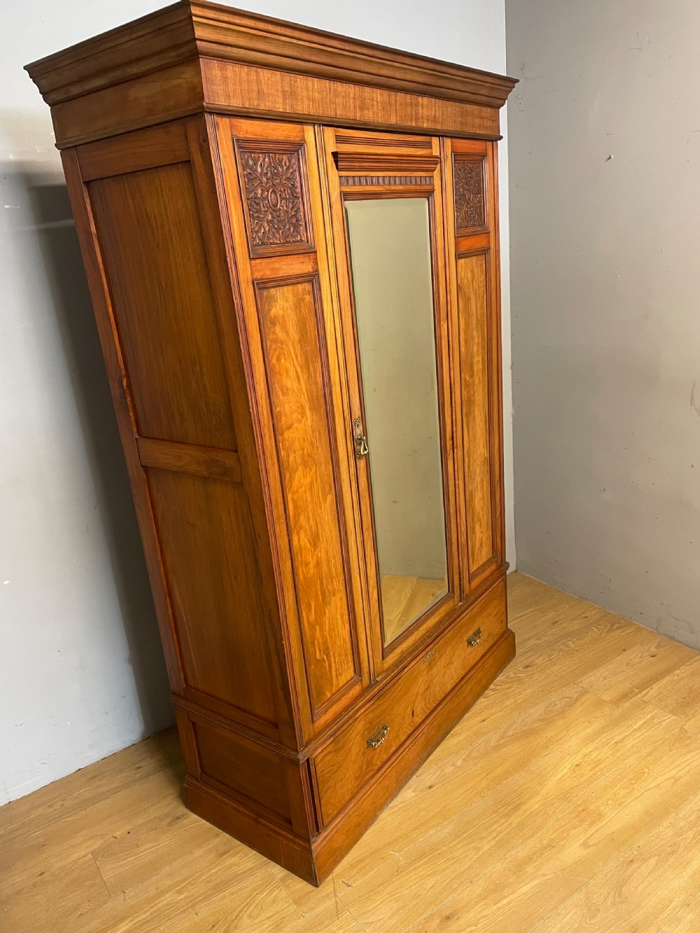 A late 19th-century Arts and Crafts satin walnut wardrobe featuring a central mirrored door, carved foliate panels, and a single base drawer. View II