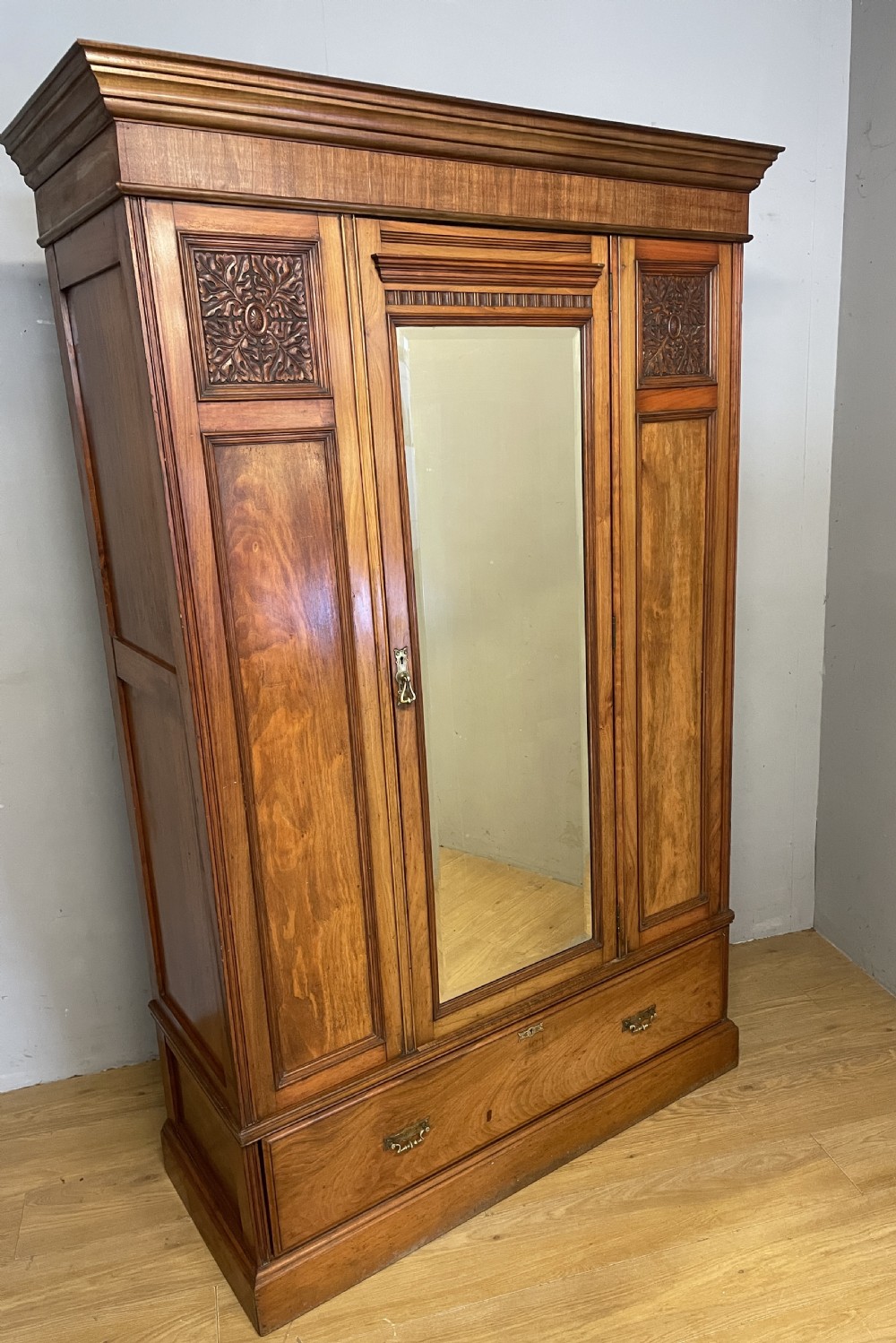 A late 19th-century Arts and Crafts satin walnut wardrobe featuring a central mirrored door, carved foliate panels, and a single base drawer. View IV