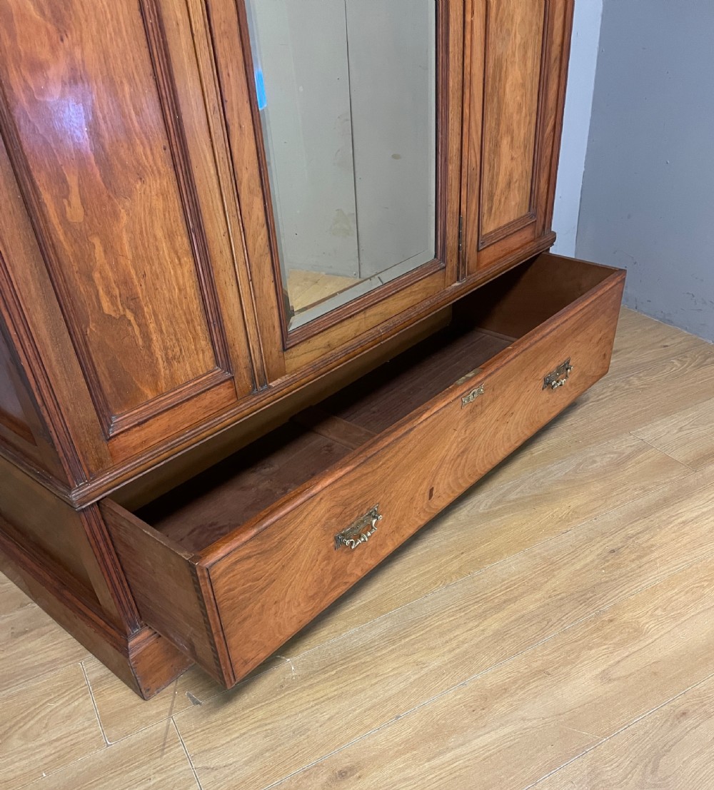 A late 19th-century Arts and Crafts satin walnut wardrobe featuring a central mirrored door, carved foliate panels, and a single base drawer. View V