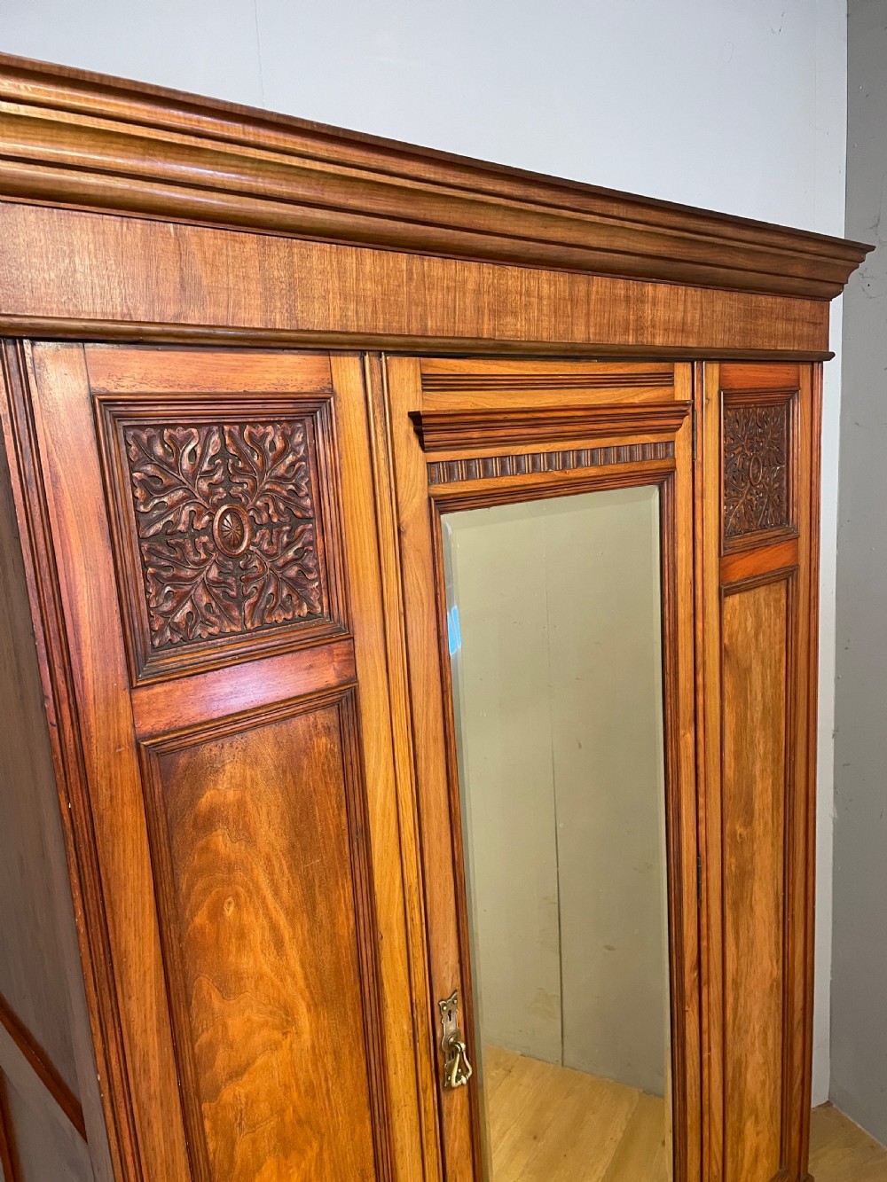 A late 19th-century Arts and Crafts satin walnut wardrobe featuring a central mirrored door, carved foliate panels, and a single base drawer. View VI