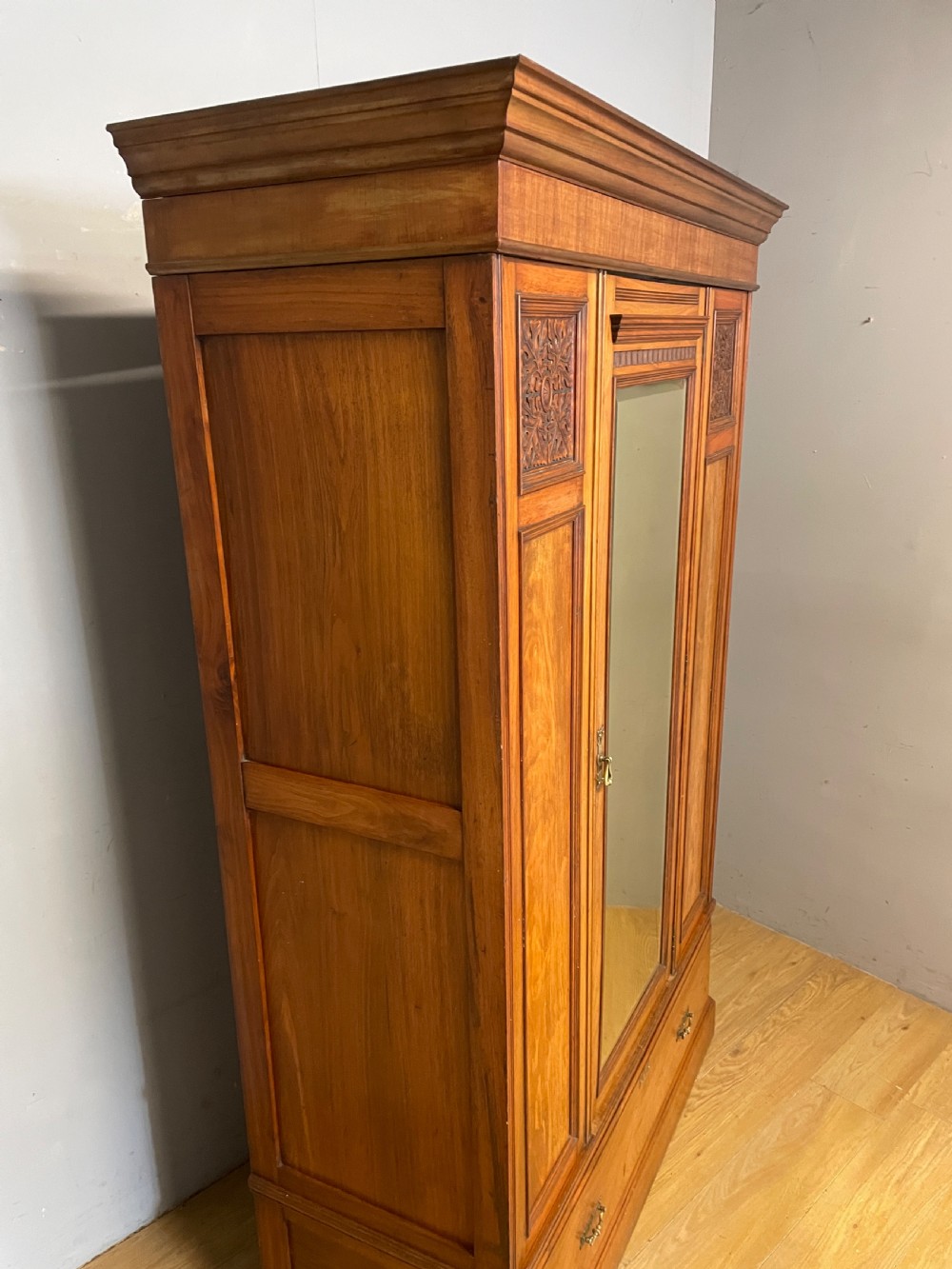 A late 19th-century Arts and Crafts satin walnut wardrobe featuring a central mirrored door, carved foliate panels, and a single base drawer. View XII