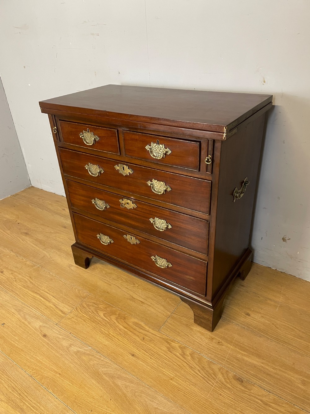 A 19th-century mahogany bachelors chest of drawers featuring a fold-over top, brass drop handles, side carrying handles, and bracket feet.