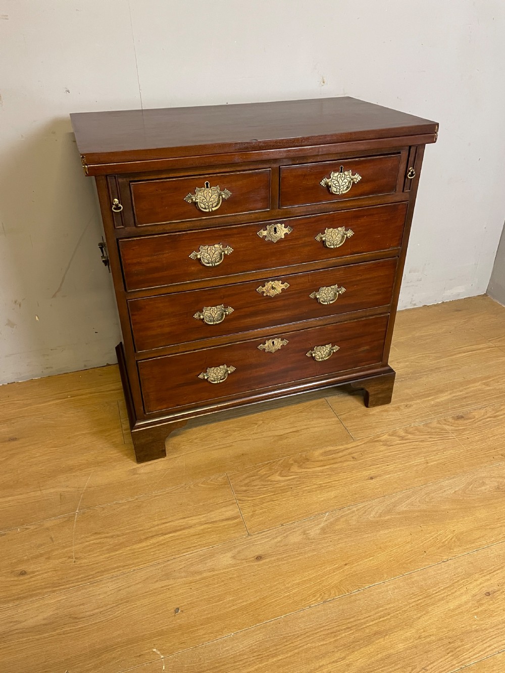 A 19th-century mahogany bachelors chest of drawers featuring a fold-over top, brass drop handles, side carrying handles, and bracket feet. View II