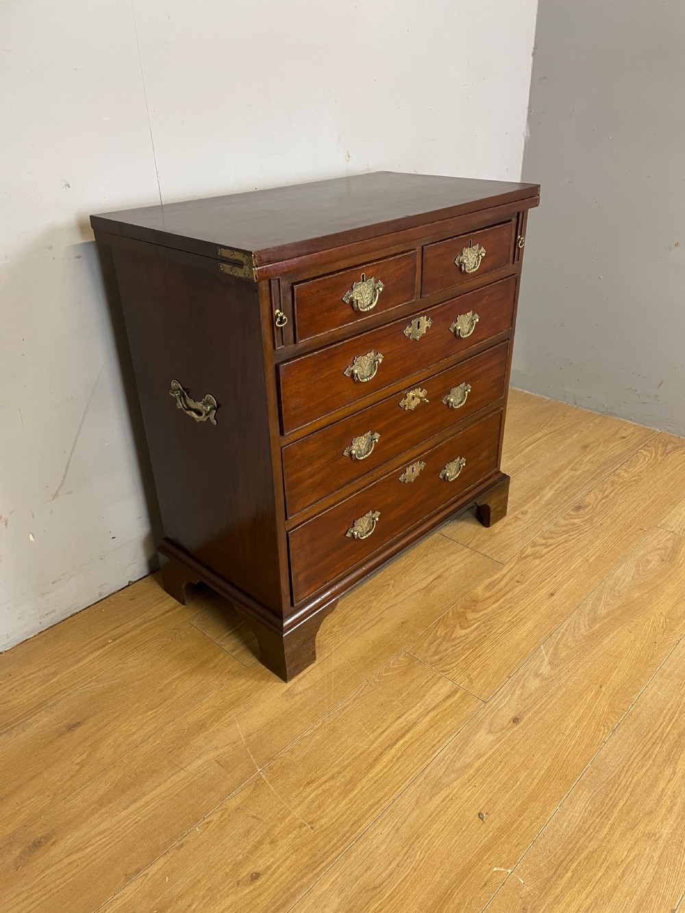 A 19th-century mahogany bachelors chest of drawers featuring a fold-over top, brass drop handles, side carrying handles, and bracket feet. View IV