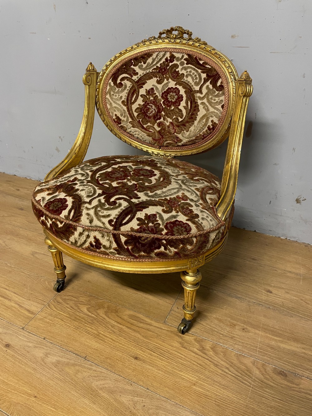A 19th-century carved giltwood nursing chair featuring patterned burgundy velvet upholstery, an oval backrest, and short turned legs with castors.