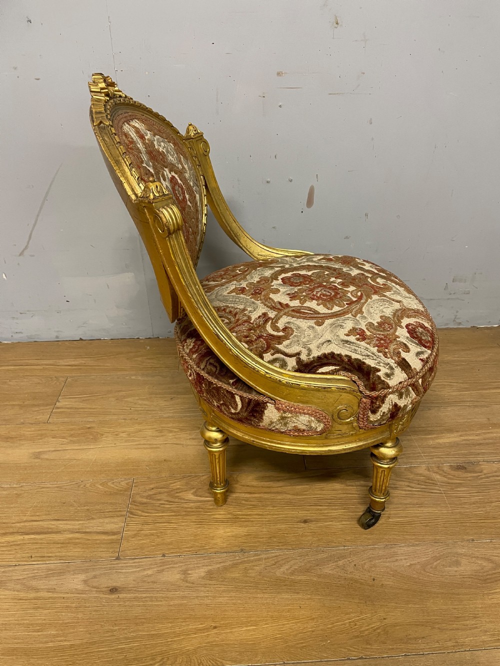 A 19th-century carved giltwood nursing chair featuring patterned burgundy velvet upholstery, an oval backrest, and short turned legs with castors. View VI