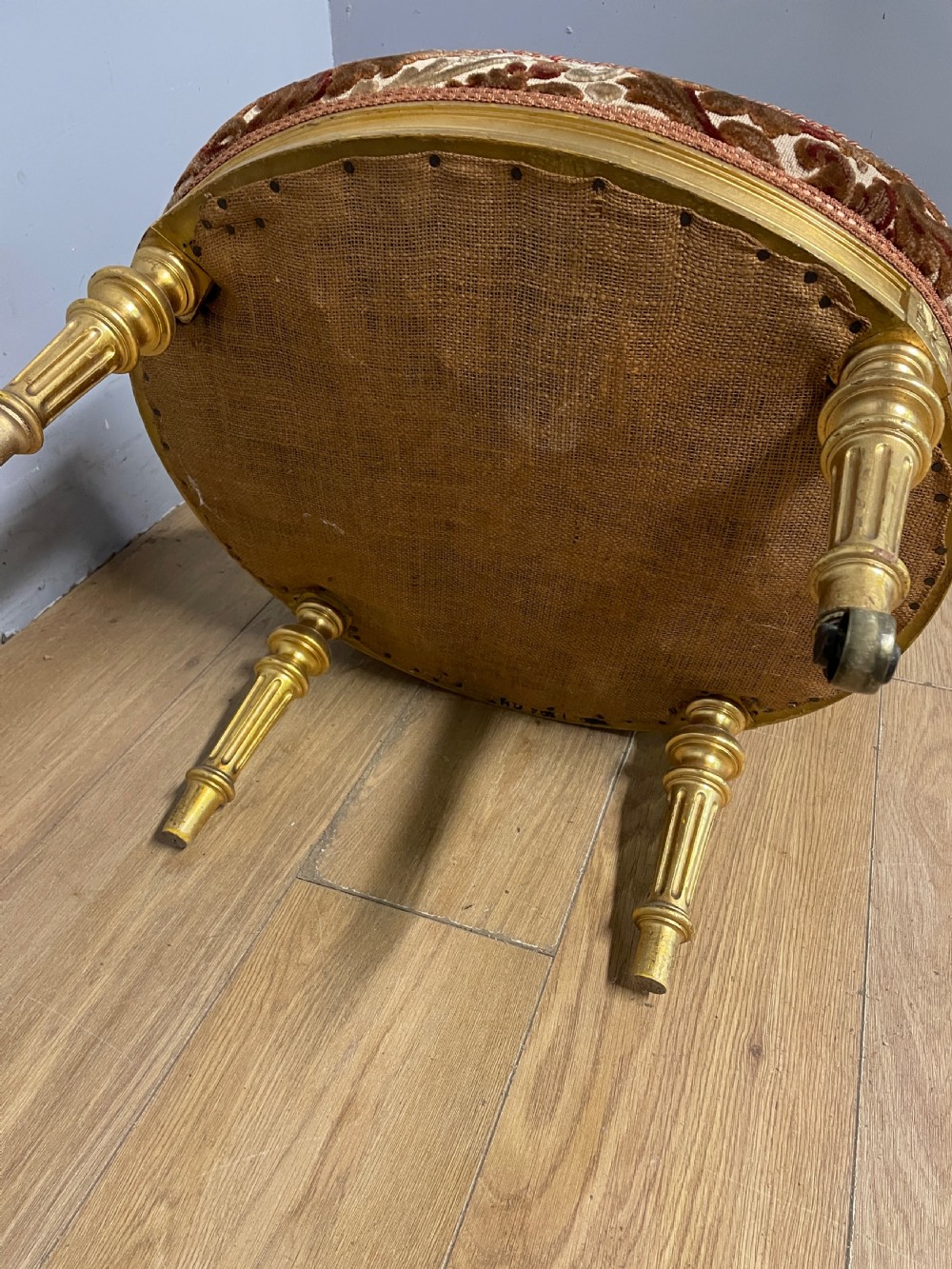 A 19th-century carved giltwood nursing chair featuring patterned burgundy velvet upholstery, an oval backrest, and short turned legs with castors. View X