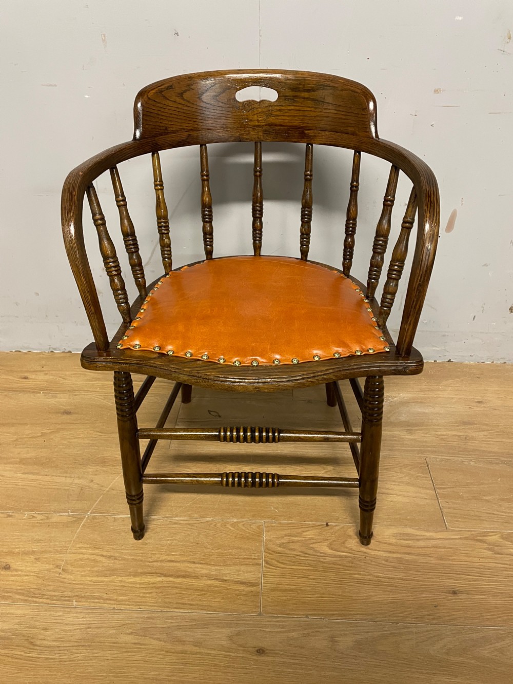An Edwardian oak and beech desk chair featuring a curved crest rail with a cut-out handle, turned spindles, and a tan leather seat secured with brass studs.