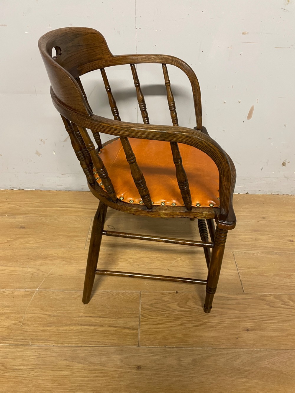 An Edwardian oak and beech desk chair featuring a curved crest rail with a cut-out handle, turned spindles, and a tan leather seat secured with brass studs. View V