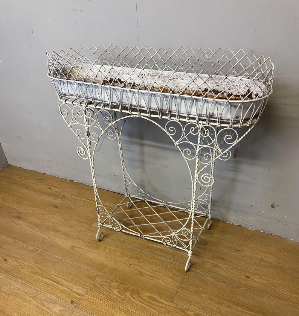 A painted distressed white Edwardian wirework plant stand featuring ornate scrollwork, a upper wire lattice basket with a zinc liner, and a lower shelf resting on scroll feet.