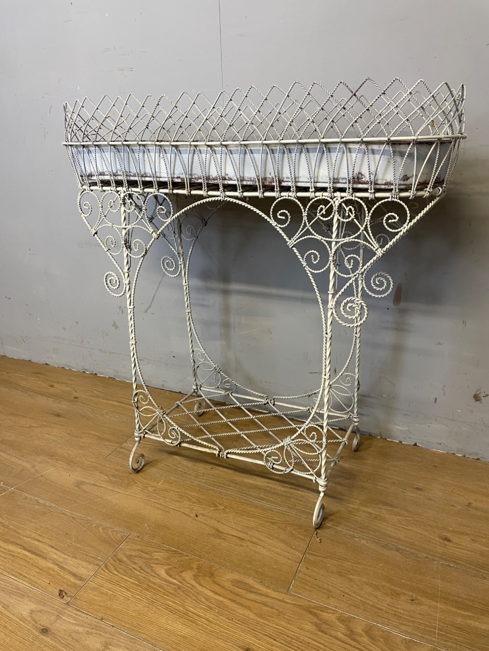 A painted distressed white Edwardian wirework plant stand featuring ornate scrollwork, a upper wire lattice basket with a zinc liner, and a lower shelf resting on scroll feet. View II