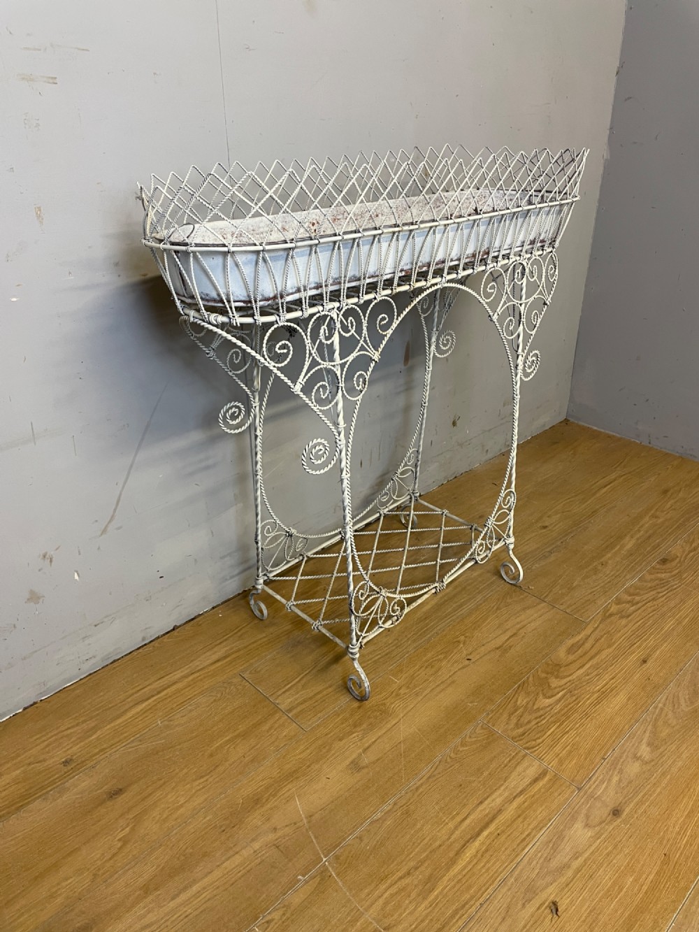 A painted distressed white Edwardian wirework plant stand featuring ornate scrollwork, a upper wire lattice basket with a zinc liner, and a lower shelf resting on scroll feet. View IV