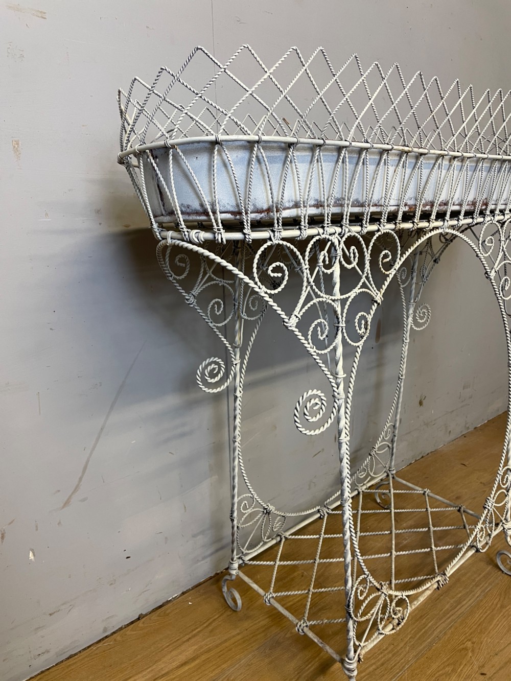 A painted distressed white Edwardian wirework plant stand featuring ornate scrollwork, a upper wire lattice basket with a zinc liner, and a lower shelf resting on scroll feet. View V