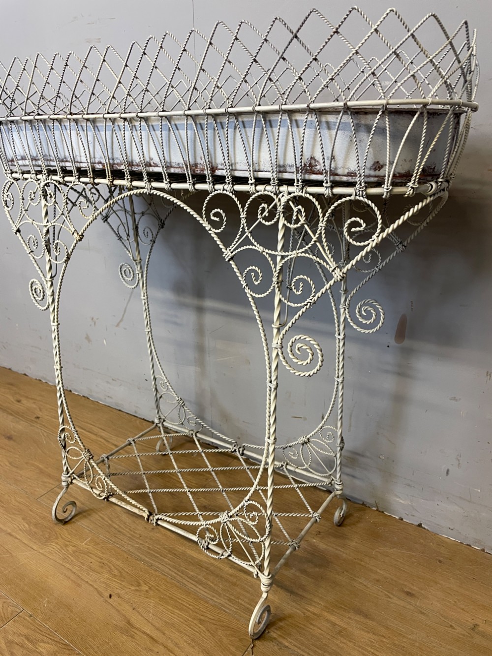A painted distressed white Edwardian wirework plant stand featuring ornate scrollwork, a upper wire lattice basket with a zinc liner, and a lower shelf resting on scroll feet. View VI