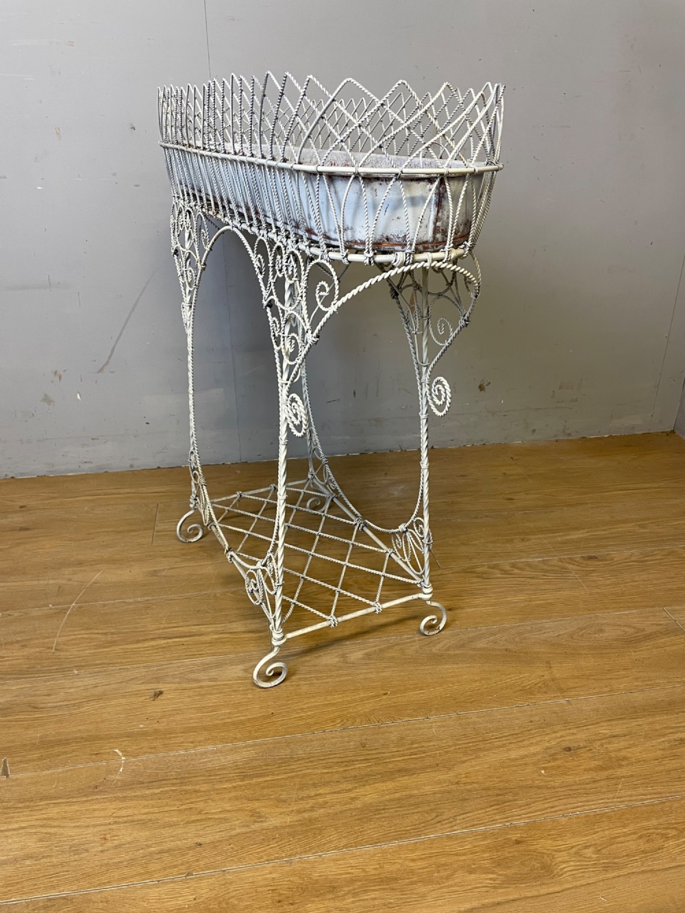 A painted distressed white Edwardian wirework plant stand featuring ornate scrollwork, a upper wire lattice basket with a zinc liner, and a lower shelf resting on scroll feet. View VII