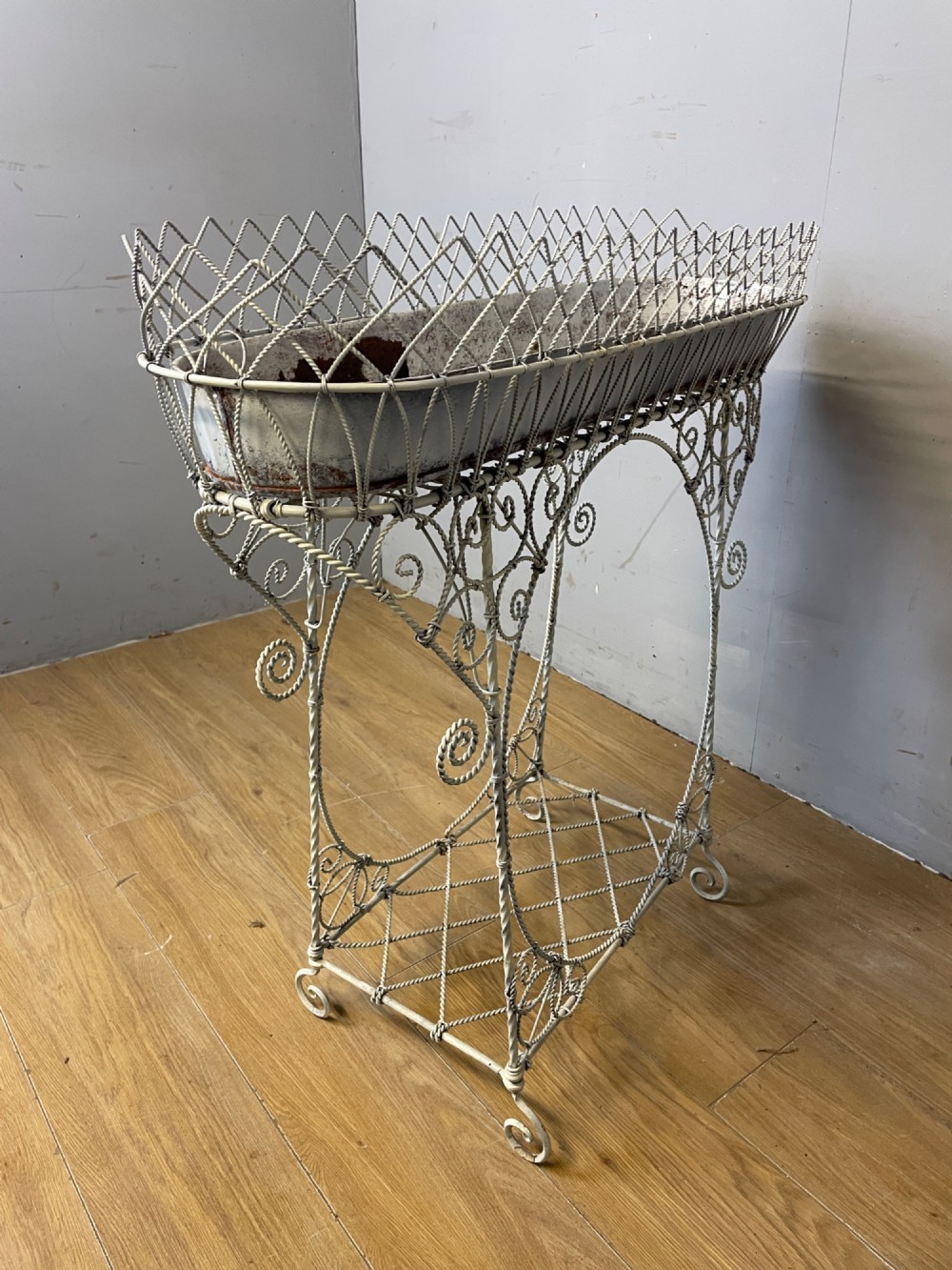 A painted distressed white Edwardian wirework plant stand featuring ornate scrollwork, a upper wire lattice basket with a zinc liner, and a lower shelf resting on scroll feet. View VIII