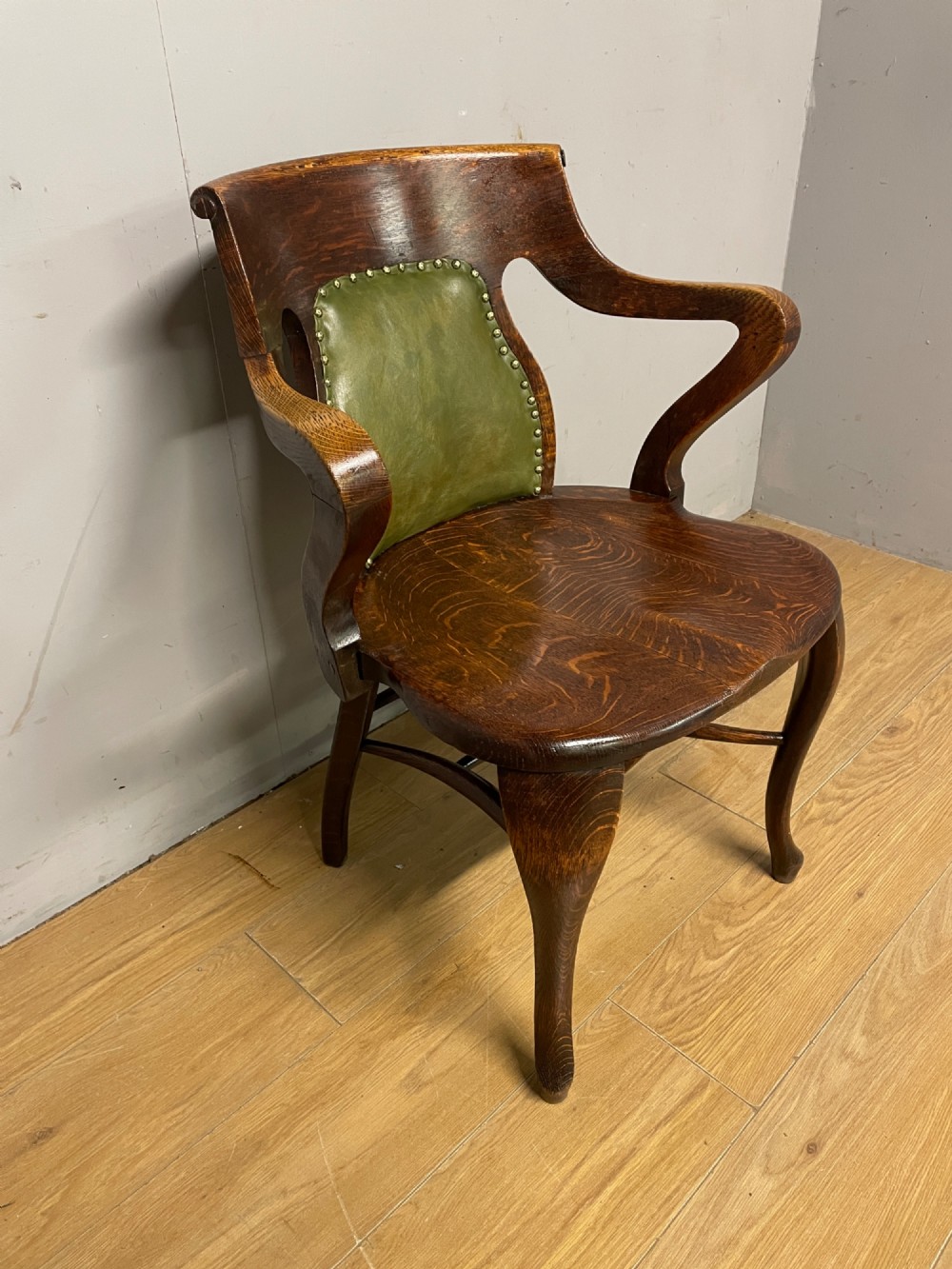 Late nineteenth-century solid oak desk chair featuring sweeping arms, a contoured timber seat, and an olive green leather back panel secured with brass studding. View IV