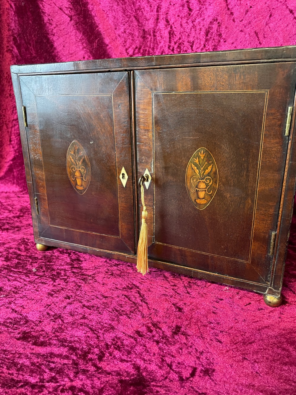 A Georgian dark mahogany tabletop cabinet featuring double doors with oval marquetry urn inlays, diamond escutcheons, and brass ball feet. View IV