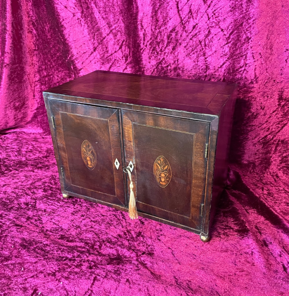 A Georgian dark mahogany tabletop cabinet featuring double doors with oval marquetry urn inlays, diamond escutcheons, and brass ball feet. View XI
