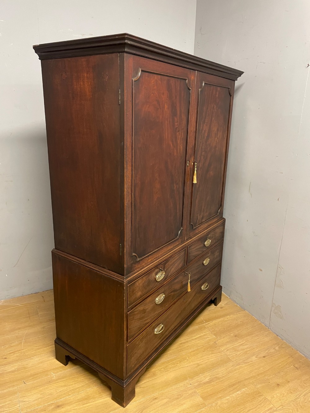 A Georgian flame mahogany linen press featuring an upper double-door cupboard with panelled doors above a lower chest of drawers with oval brass handles. View IX