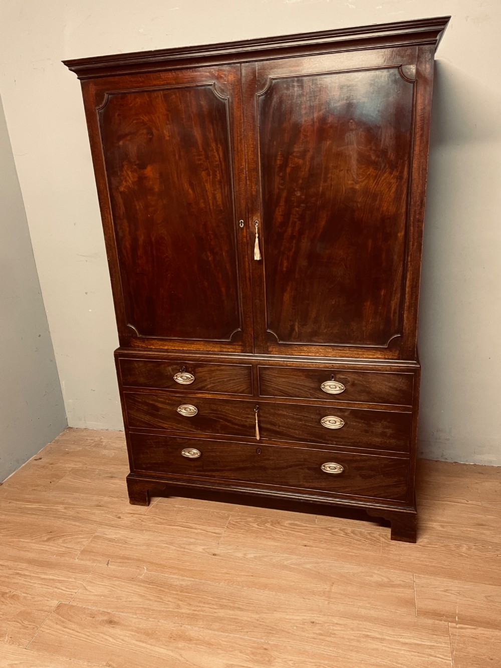 A Georgian flame mahogany linen press featuring an upper double-door cupboard with panelled doors above a lower chest of drawers with oval brass handles. View X