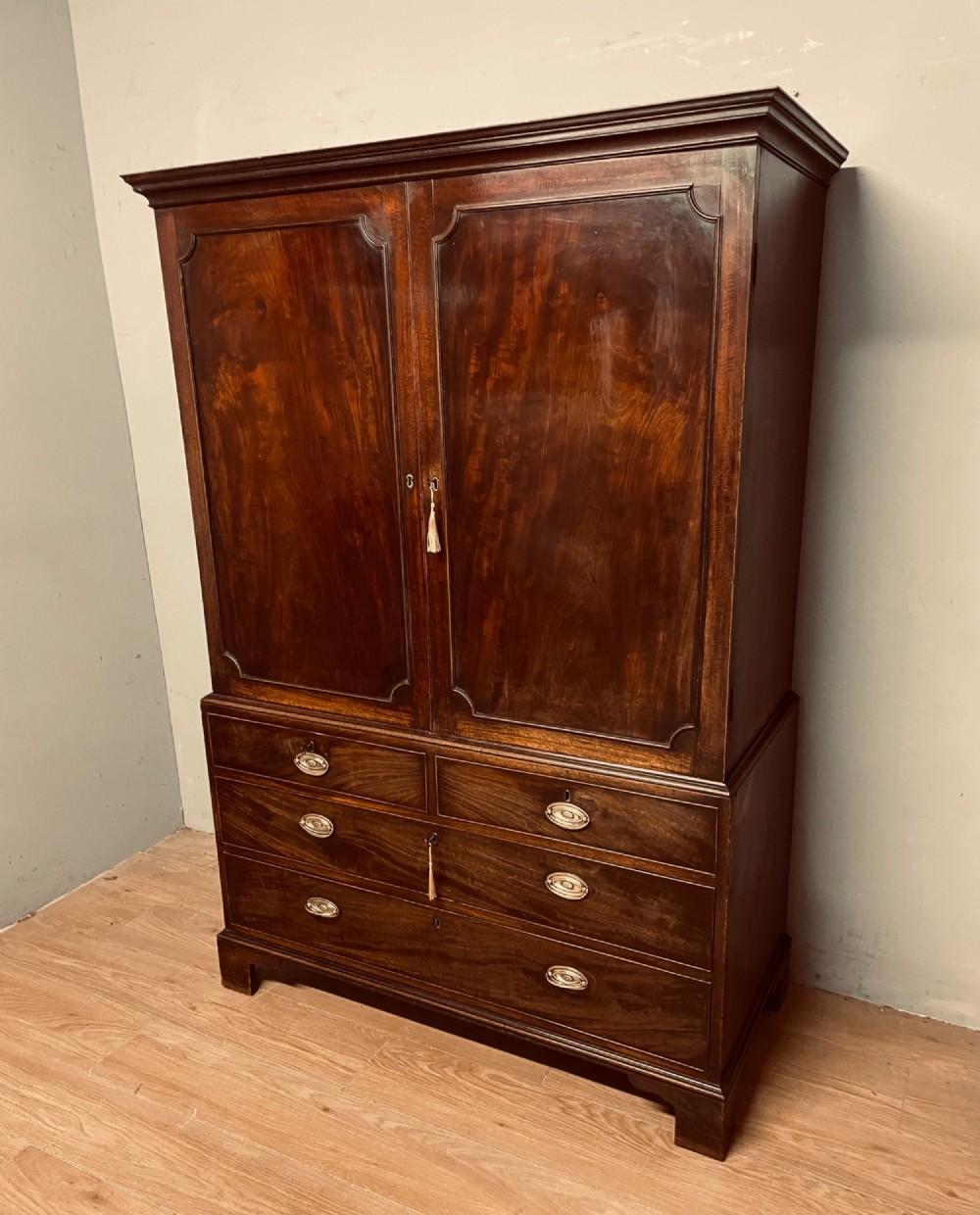 A Georgian flame mahogany linen press featuring an upper double-door cupboard with panelled doors above a lower chest of drawers with oval brass handles. View XI