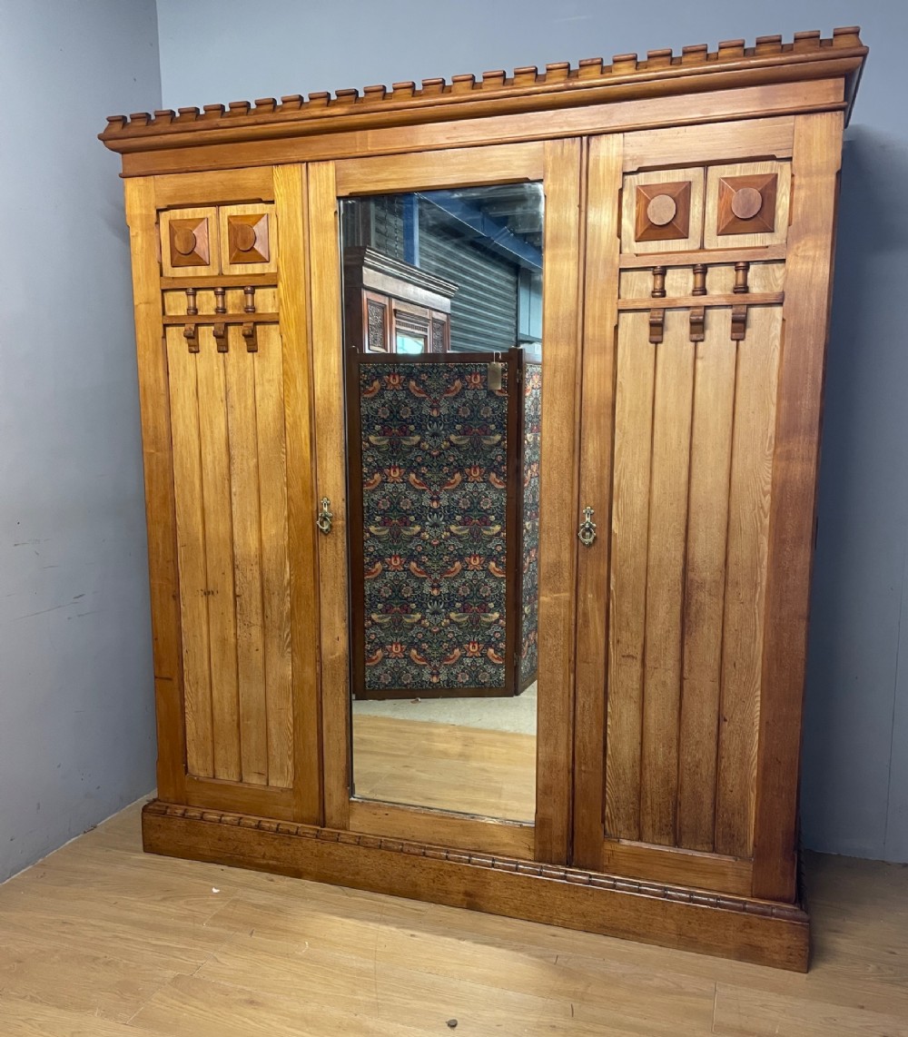 A large golden ash Arts and Crafts triple wardrobe featuring a central mirrored door flanked by panelled wooden doors with castellated cornice detailing.