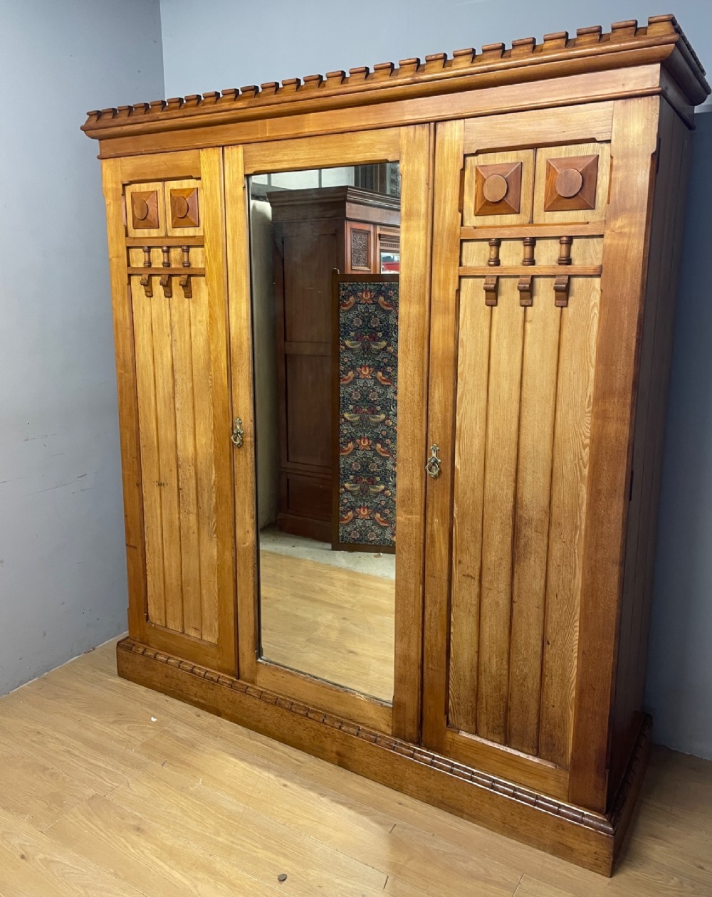 A large golden ash Arts and Crafts triple wardrobe featuring a central mirrored door flanked by panelled wooden doors with castellated cornice detailing. View II