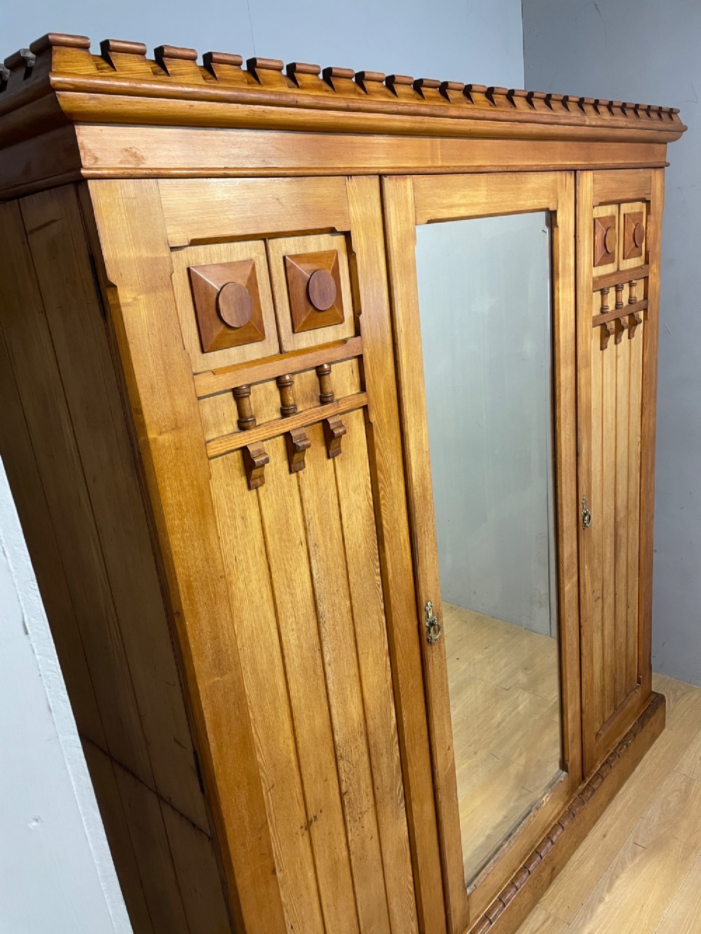 A large golden ash Arts and Crafts triple wardrobe featuring a central mirrored door flanked by panelled wooden doors with castellated cornice detailing. View IV