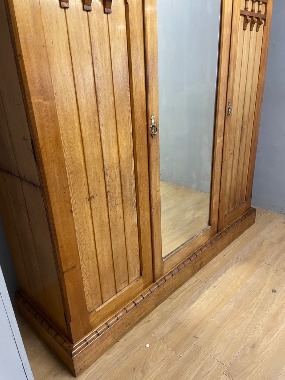 A large golden ash Arts and Crafts triple wardrobe featuring a central mirrored door flanked by panelled wooden doors with castellated cornice detailing. View V