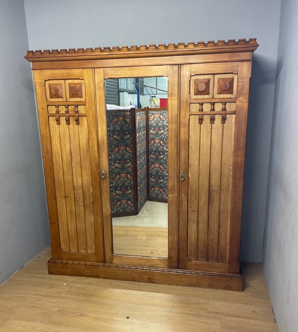 A large golden ash Arts and Crafts triple wardrobe featuring a central mirrored door flanked by panelled wooden doors with castellated cornice detailing. View XII