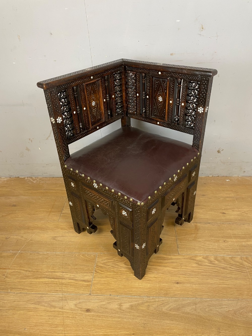 A 19th-century carved dark wood corner chair featuring mother-of-pearl floral inlay, detailed Mashrabiya panels, and a brown leather seat secured with brass studs.