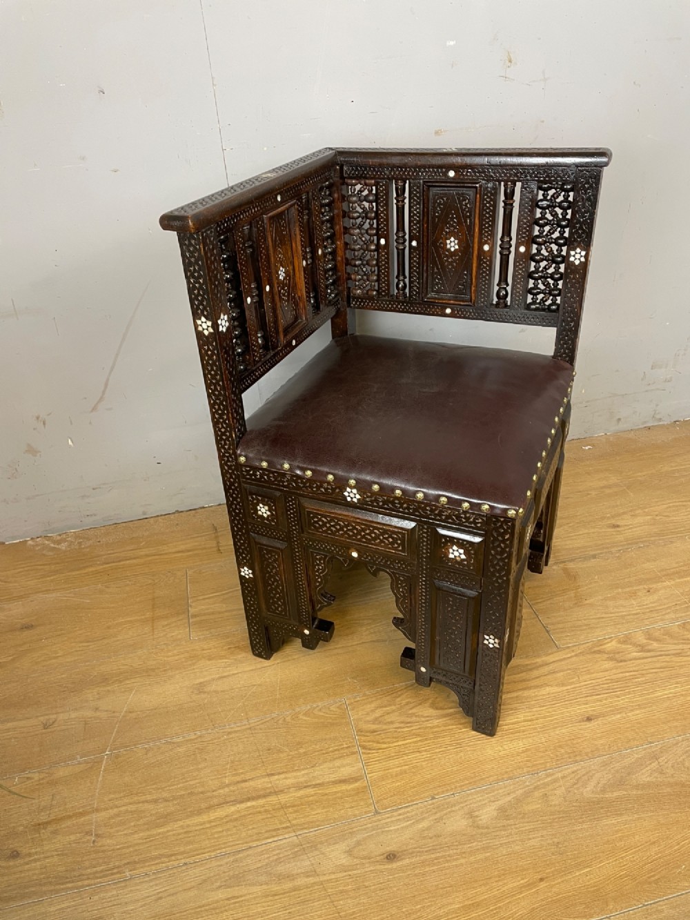 A 19th-century carved dark wood corner chair featuring mother-of-pearl floral inlay, detailed Mashrabiya panels, and a brown leather seat secured with brass studs. View II