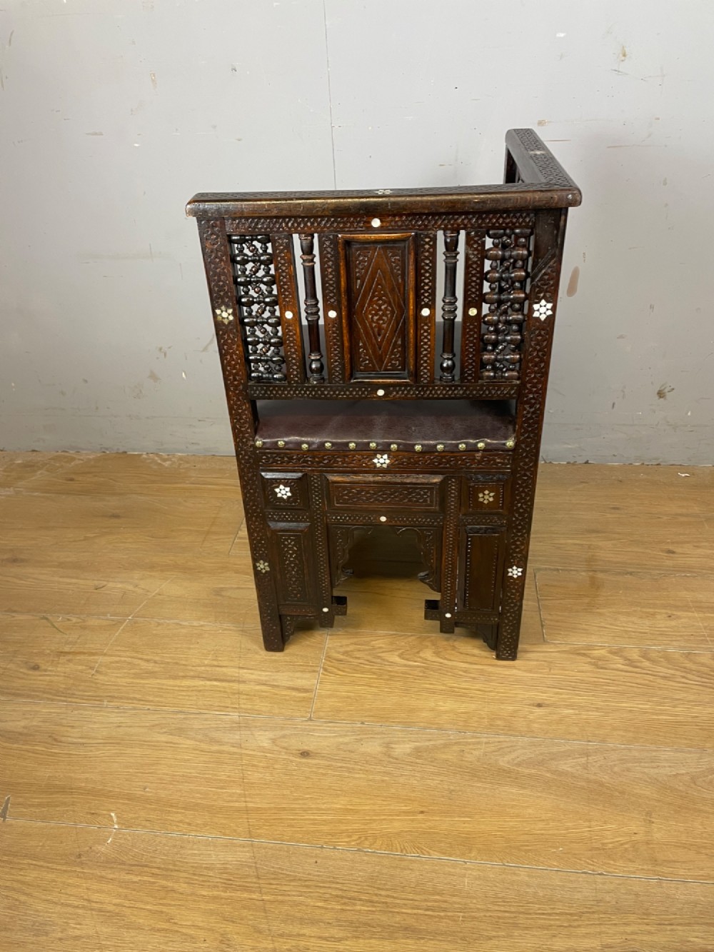 A 19th-century carved dark wood corner chair featuring mother-of-pearl floral inlay, detailed Mashrabiya panels, and a brown leather seat secured with brass studs. View V